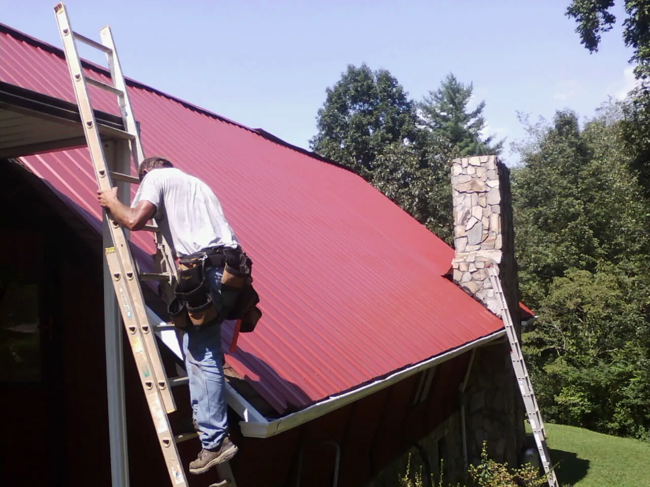 Roofer on a ladder beside a red metal roof and stone chimney — Soffit & Fascia Repair by Roofing Brothers in Plantsville